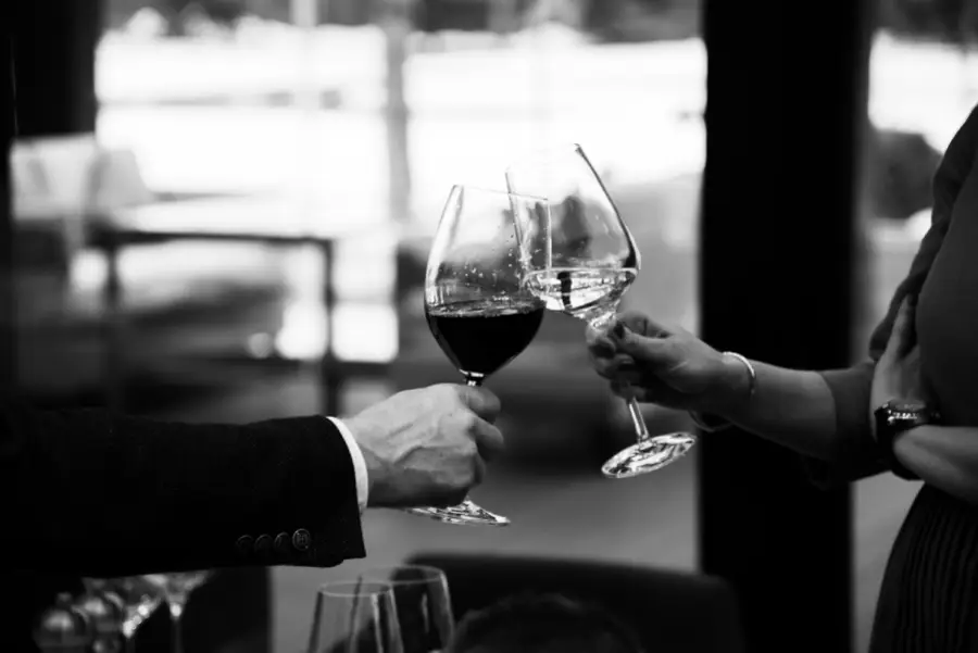 Two people toasting with wine glasses at a dining table in a black-and-white setting.